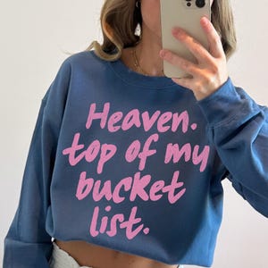 May include: A cropped, blue sweatshirt with the phrase "Heaven. top of my bucket list." printed in pink. The sweatshirt has long sleeves and a crew neck. The image is taken from a medium distance.