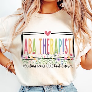 May include: Cream-colored t-shirt with the words "ABA THERAPIST" in colorful letters inside a black rectangle. Below the text is a floral design and the words "planting seeds that last forever."