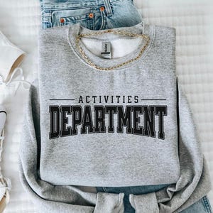 Peut inclure: Un sweat-shirt gris chiné avec l'inscription "ACTIVITIES DEPARTMENT" en lettres noires. Un collier chaîne doré est posé sur le sweat-shirt. Des baskets blanches et un jean bleu sont visibles en arrière-plan.