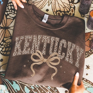 Peut inclure: Un sweat-shirt marron avec le mot "KENTUCKY" en imprimé léopard. Un motif de nœud beige est centré en dessous. Le sweat-shirt est tenu par des mains, avec un tissu à motifs et des bougies en arrière-plan.