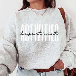 May include: A light gray heathered crewneck sweatshirt featuring the word ACTIVITIES in large white block letters with the word department written in a black cursive script layered horizontally across the center of the white text.