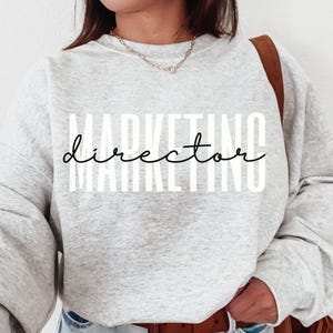 Op de afbeelding: Een lichtgrijs sweatshirt met de tekst "MARKETING director" gedrukt in een mix van vette en cursieve lettertypen. De longsleeve wordt gedragen met een blauwe jeans en een bruine riem. Een bruine schoudertas is ook zichtbaar.