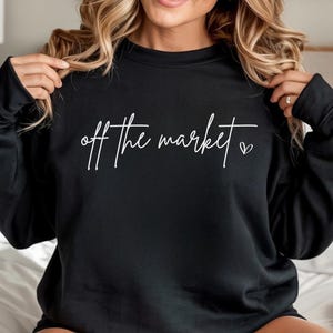 Puede incluir: Sudadera negra con la frase "off the market" en escritura blanca y un pequeño corazón. La sudadera es de estilo casual.