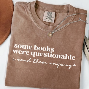 May include: A brown Comfort Colors t-shirt with the text "some books were questionable i read them anyway" in white. A silver necklace with a small pendant is draped across the shirt. The shirt is folded on a white surface.