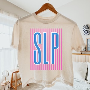 May include: A cream-colored t-shirt with the letters "SLP" in blue, outlined in white, centered on the front. The letters are set against a background of pink vertical stripes. The shirt is hanging on a wooden hanger.