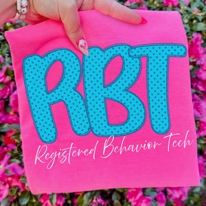 May include: A bright pink t-shirt featuring the large teal letters RBT with a dark teal polka dot pattern. Below the letters, the text Registered Behavior Tech is printed in a white cursive font. The shirt is displayed against a floral background.