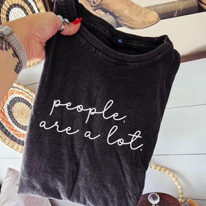 May include: A charcoal grey short-sleeve t-shirt with a vintage-style washed finish. The front features the white cursive text "people, are a lot." The shirt has a ribbed crew neckline and a black interior neck label with the letter R printed on it.