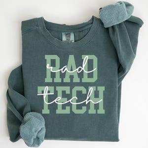 May include: A dark teal sweatshirt with the words "RAD TECH" in a light green block font and white cursive. The sweatshirt has a crew neck and long sleeves with folded cuffs. The Comfort Colors brand tag is visible.
