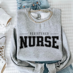 May include: A grey sweatshirt displaying the text "REGISTERED NURSE" in black, bold lettering. A gold chain necklace is placed on the sweatshirt, alongside blue jeans and white trainers.