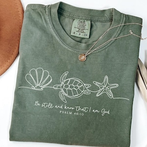 Puede incluir: Camiseta verde oliva con un dibujo lineal blanco de una concha, una tortuga y una estrella de mar. El texto "Be still and know that I am God, Psalm 46:10" está impreso debajo. Un collar dorado con un colgante descansa sobre la camiseta.