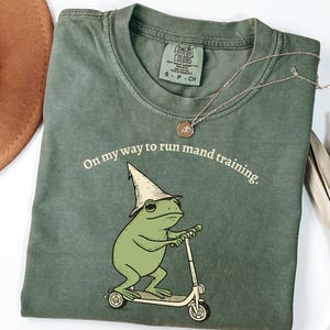 Könnte beinhalten: Grünes Comfort Colors T-Shirt mit einem Frosch auf einem Roller und einem Zaubererhut. Der Text "On my way to run mand training" ist über dem Frosch aufgedruckt. Eine goldene Halskette liegt auf dem Shirt.