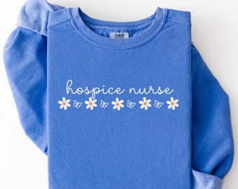 May include: A blue sweatshirt with the words "hospice nurse" in white script, decorated with white daisies and butterflies. The sweatshirt is folded, with the sleeves visible. The Comfort Colors brand tag is visible.