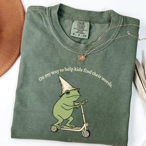 May include: An olive green t-shirt with the text "On my way to help kids find their words." features a cartoon frog riding a scooter and wearing a wizard hat. A gold necklace with a pendant is also visible.