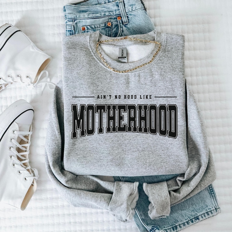 May include: A light gray sweatshirt with the text "AIN'T NO HOOD LIKE MOTHERHOOD" in black block letters. The sweatshirt is paired with blue jeans, a gold chain necklace, and white high-top sneakers.