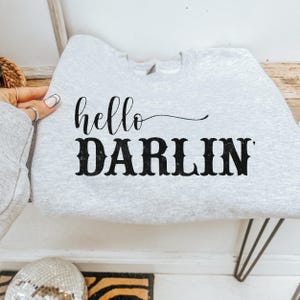 May include: A light gray sweatshirt with the words "hello DARLIN" printed in black. The word "hello" is in a cursive font, and "DARLIN" is in a bold, block font. The sweatshirt is folded.