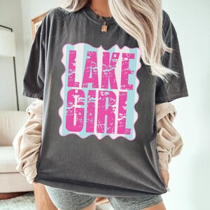 Puede incluir: Camiseta gris oscuro con el texto "LAKE GIRL" en rosa desgastado, sobre un fondo a rayas azul claro y blanco. El dise&ntilde;o est&aacute; enmarcado por un borde festoneado.