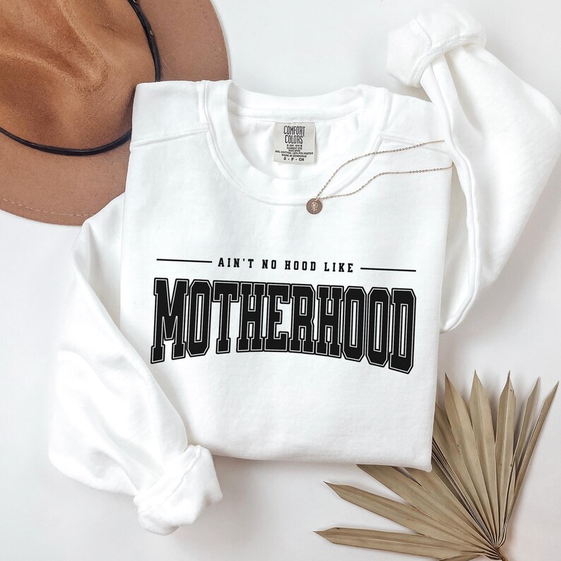 May include: White crew neck sweatshirt with the text "AIN'T NO HOOD LIKE MOTHERHOOD" in black block letters. A gold necklace and a brown hat are also visible. The sweatshirt is made of a soft material.