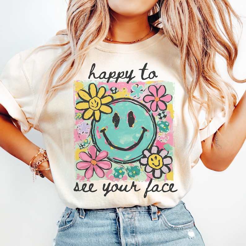 May include: Cream-coloured t-shirt with a colourful graphic featuring a turquoise smiley face surrounded by flowers. The text "happy to see your face" is above and below the graphic. The shirt has short sleeves.