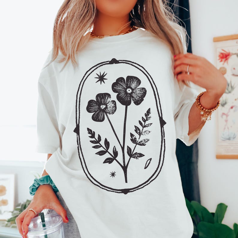 May include: White t-shirt featuring a black floral design within an oval frame. The design includes two flowers, leaves, and stars. The shirt has short sleeves and a relaxed fit, suitable for casual wear.