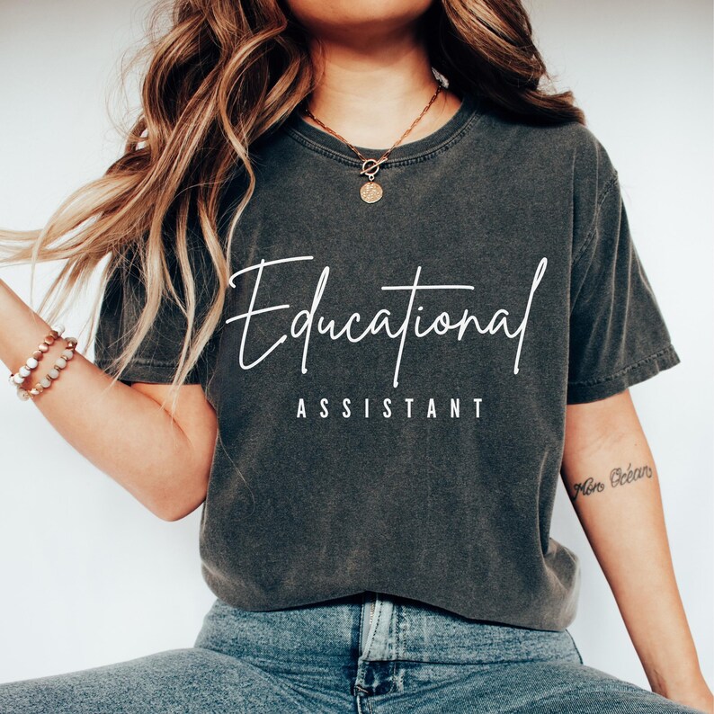 May include: Dark gray t-shirt with the words "Educational Assistant" in white script and block letters. The shirt is a crew neck and has a slightly faded appearance. The overall style is casual and comfortable.
