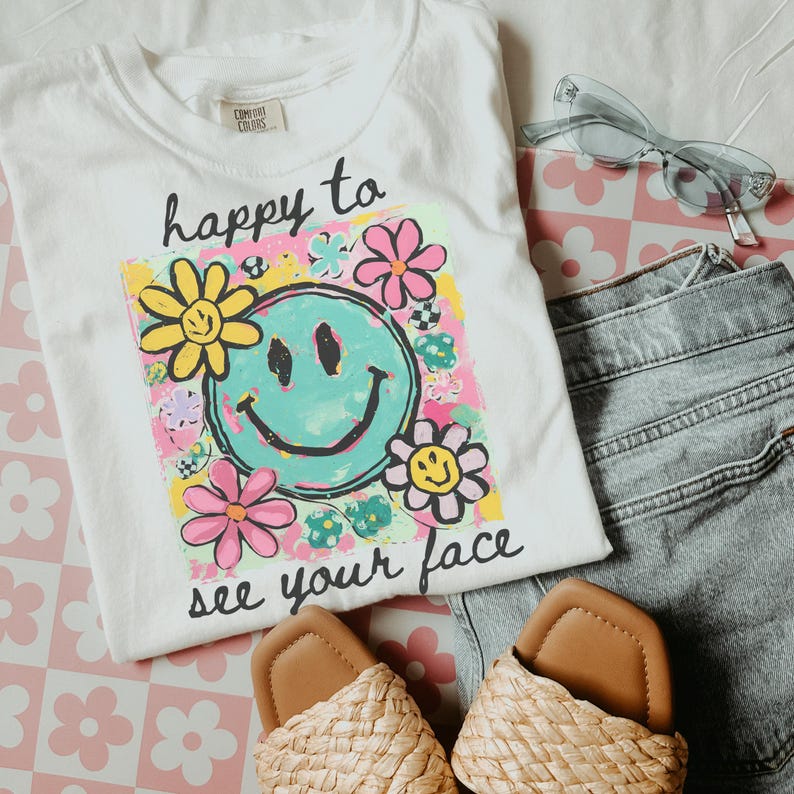 May include: A white t-shirt featuring the phrase "happy to see your face" and a vibrant smiley face design. The image includes denim shorts, woven sandals, and light blue sunglasses. The items are arranged on a pink floral patterned surface.