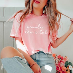 May include: A short-sleeved, crew-neck t-shirt in a soft, washed-out coral pink color. The front of the shirt features the white, cursive-style text "people. are a lot." printed across the chest area. The shirt is paired with light blue denim jeans.