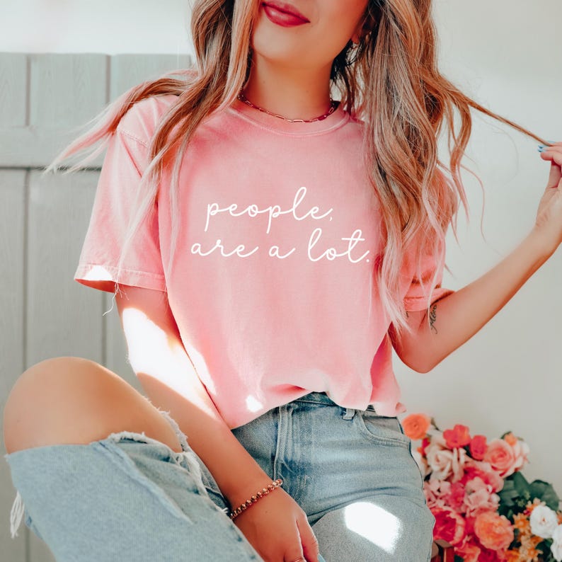 May include: A short-sleeved, crew-neck t-shirt in a soft, washed-out coral pink color. The front of the shirt features the white, cursive-style text "people. are a lot." printed across the chest area. The shirt is paired with light blue denim jeans.