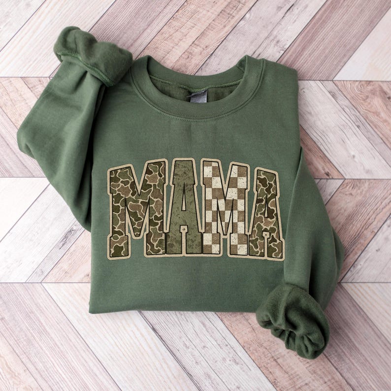 Op de afbeelding: Een olijfgroene sweatshirt met het woord "MAMA" in blokletters. De letters zijn versierd met een camouflagepatroon en een dambordpatroon. De mouwen zijn gerimpeld.