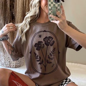 May include: A brown t-shirt featuring a floral design within an oval frame. The graphic includes two flowers, leaves, and stars. The shirt has short sleeves and a relaxed fit, suitable for casual wear.