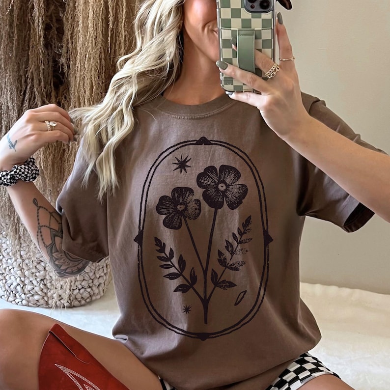 May include: A brown t-shirt featuring a floral design within an oval frame. The graphic includes two flowers, leaves, and stars. The shirt has short sleeves and a relaxed fit, suitable for casual wear.