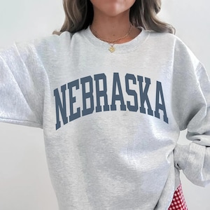 May include: A light gray sweatshirt with the word "NEBRASKA" printed in navy blue block letters across the chest. The sweatshirt has long sleeves and a crew neck. The model is wearing a gold necklace.