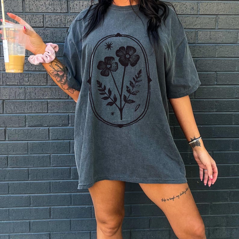 May include: Dark gray oversized t-shirt featuring a floral design within an oval frame. The graphic includes two flowers, leaves, and stars. The shirt has short sleeves and a relaxed fit, suitable for casual wear.