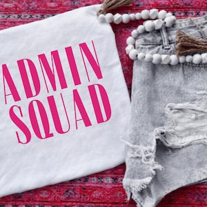 May include: White t-shirt with the text "ADMIN SQUAD" in bright pink. The shirt is paired with distressed denim shorts and a beaded garland. The items are arranged on a patterned red surface.