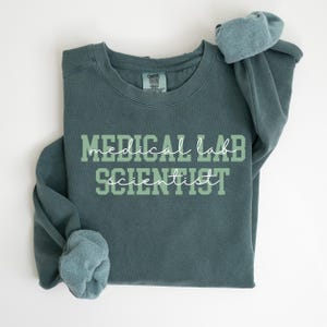 May include: A teal-colored sweatshirt with the words "MEDICAL LAB SCIENTIST" printed in a two-tone green font. The sleeves are folded, and the garment has a crew neck. The sweatshirt is made of a soft material.
