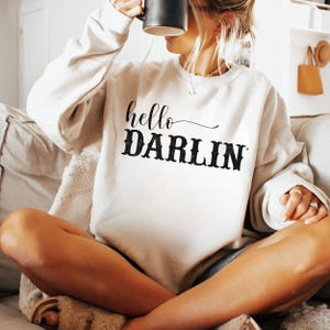 May include: Cream-colored sweatshirt with the text "hello DARLIN" in black script and block letters. The sweatshirt is long-sleeved and appears to be made of a soft material. The person is holding a black mug.