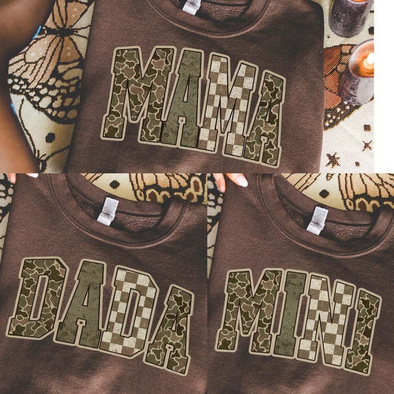 Op de afbeelding: Drie bruine sweatshirts met de woorden "MAMA", "DADA" en "MINI" erop gedrukt. De letters hebben een camouflage- en dambordpatroon. De sweatshirts worden gepresenteerd op een oppervlak met een patroon.