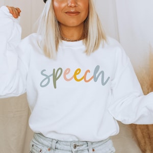May include: A white sweatshirt with the word "speech" in pastel colors across the front. The word is written in a cursive font with each letter a different color: green, pink, yellow, and blue. The sweatshirt has long sleeves.