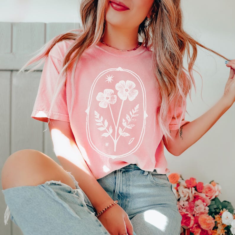 May include: A coral pink short-sleeved t-shirt featuring a white graphic print of two flowers and leafy stems enclosed within an oval frame. The design includes small star-like accents above and below the floral illustration on the front chest.
