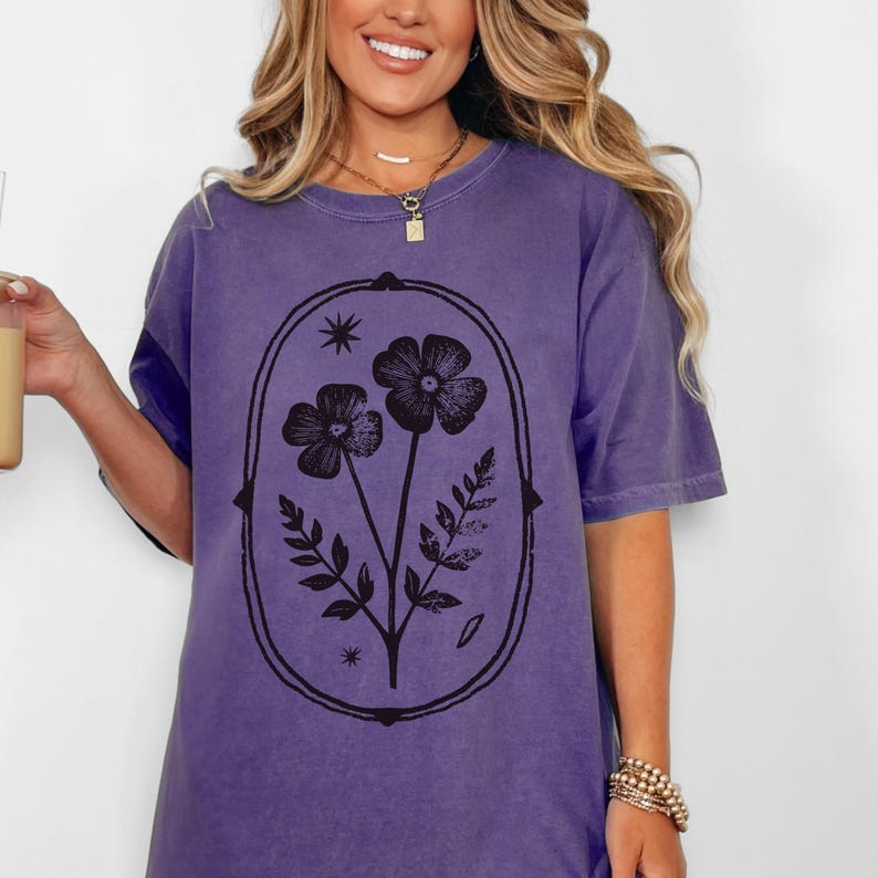 May include: A purple t-shirt featuring a black floral design within an oval frame. The design includes two flowers, leaves, and stars. The shirt has short sleeves and a relaxed fit.