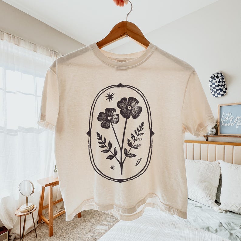 May include: Cream-colored t-shirt with a black floral design. The graphic features two flowers, leaves, and stars within an oval frame. The shirt is hanging on a wooden hanger.