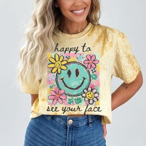 May include: Yellow t-shirt with a graphic of a smiling face in a square frame, surrounded by colourful flowers. The text "happy to see your face" is above and below the graphic. The shirt has a tie-dye effect.