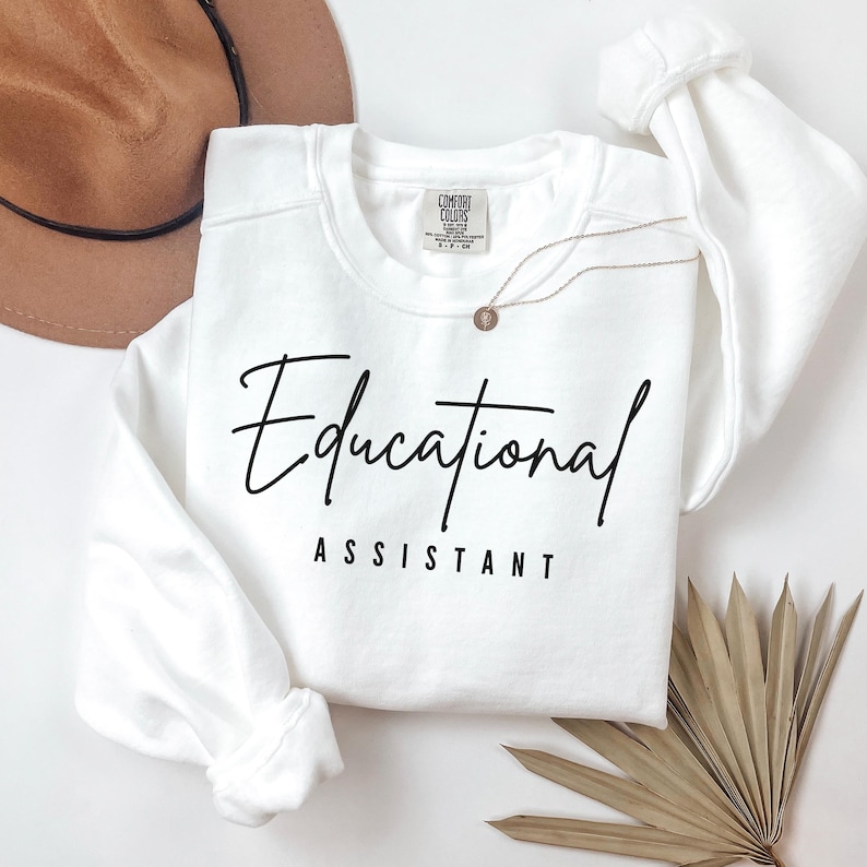 May include: White crewneck sweatshirt with the words "Educational Assistant" in black script. A gold necklace and a brown hat are also visible. The sweatshirt is made of a soft material.