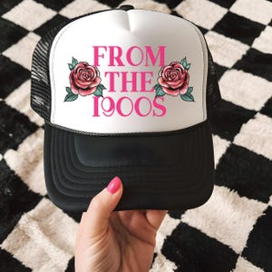 May include: Black and white trucker hat with a white front panel. The panel displays the pink text "FROM THE 1900S" and pink roses with green leaves. The hat has a black brim and a mesh back.