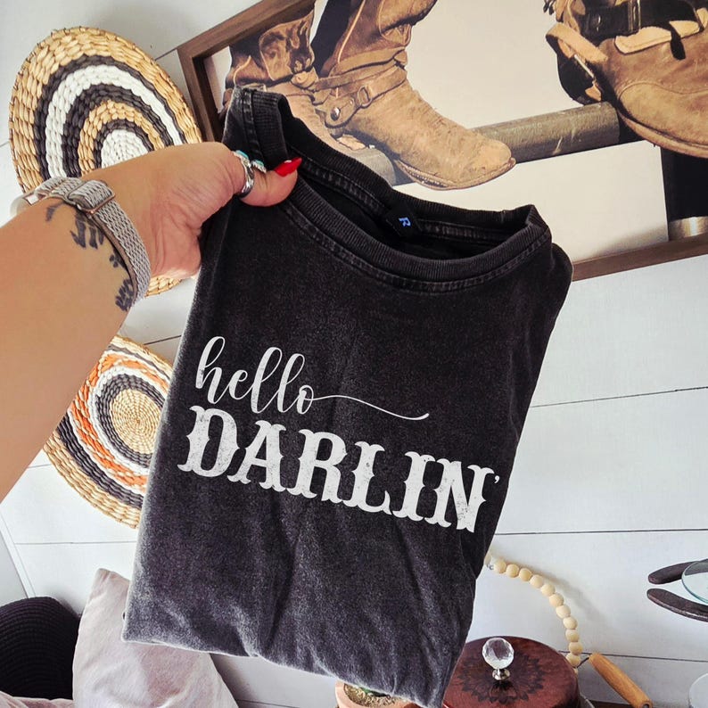 May include: A dark gray t-shirt with the words "hello DARLIN" in white script. The shirt is folded and held up against a white-paneled wall with decorative items in the background. The shirt has a vintage, worn look.