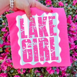 Puede incluir: Una camiseta rosa brillante con las palabras "LAKE GIRL" en letras rosas desgastadas. El texto est&aacute; sobre un fondo a rayas blancas y celeste con un borde festoneado. La camiseta est&aacute; sujeta por una mano.