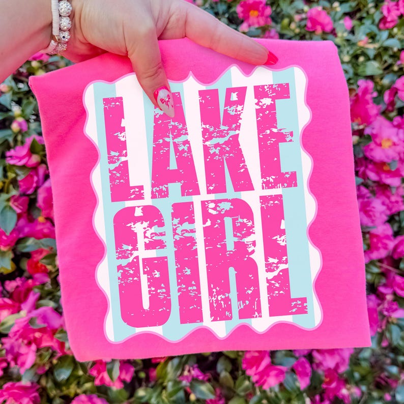 Puede incluir: Una camiseta rosa brillante con las palabras "LAKE GIRL" en letras rosas desgastadas. El texto est&aacute; sobre un fondo a rayas blancas y celeste con un borde festoneado. La camiseta est&aacute; sujeta por una mano.