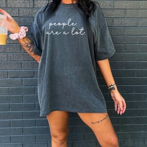 May include: A charcoal grey short-sleeve t-shirt featuring the white cursive text "people. are a lot." printed across the chest. The garment has a relaxed, oversized fit with a crew neckline and a washed, vintage-inspired fabric texture.