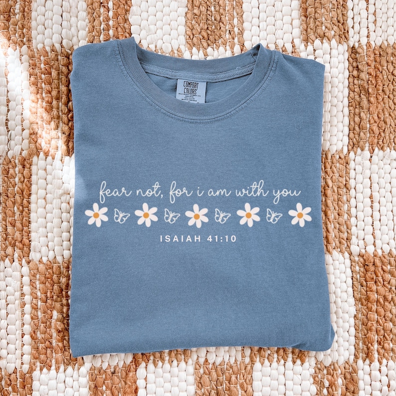 May include: A blue Comfort Colors t-shirt with the text "fear not, for i am with you" in white script. The text is accompanied by a row of white daisies and butterflies. The text "ISAIAH 41:10" is printed below the quote.