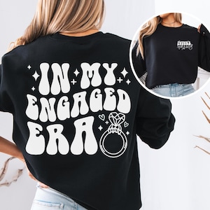 May include: Black sweatshirt with the text "IN MY ENGAGED ERA" in white, along with a diamond ring graphic. The design includes stars and hearts. A smaller image shows the text "FUTURE MRS" on the sleeve.