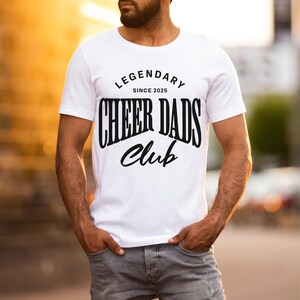 May include: White t-shirt with black text that reads "Legendary Since 2025 Cheer Dads Club".